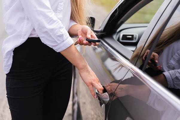 A woman is holding her phone while standing next to the door of a car.